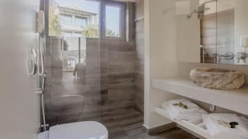 Un baño moderno con ducha envuelta en vidrio, inodoro blanco, lavamanos de piedra y toallas dobladas ordenadamente en un estante.
