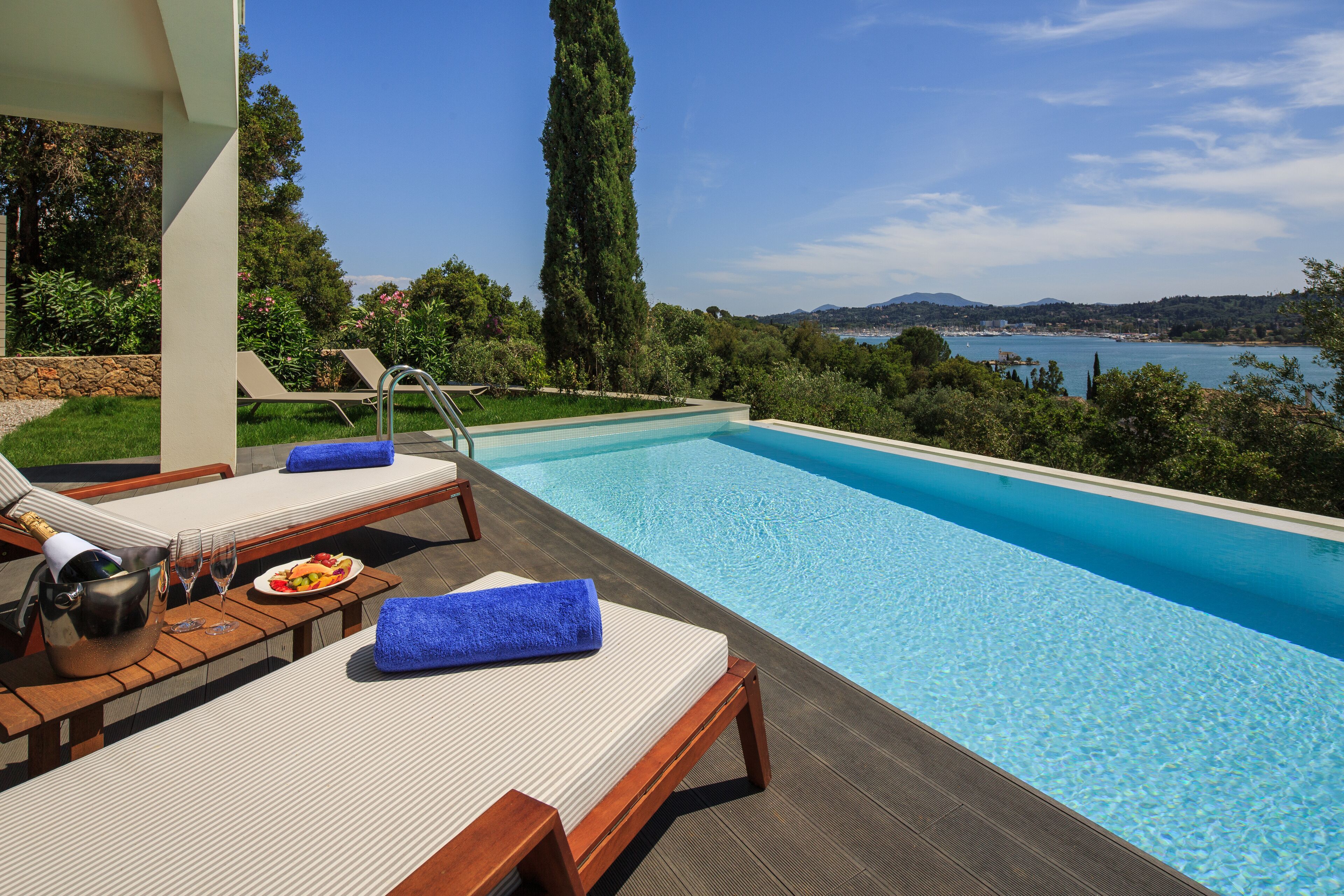 A poolside area with lounge chairs, a table with a bottle and glasses, and a view of the sea and hills.
