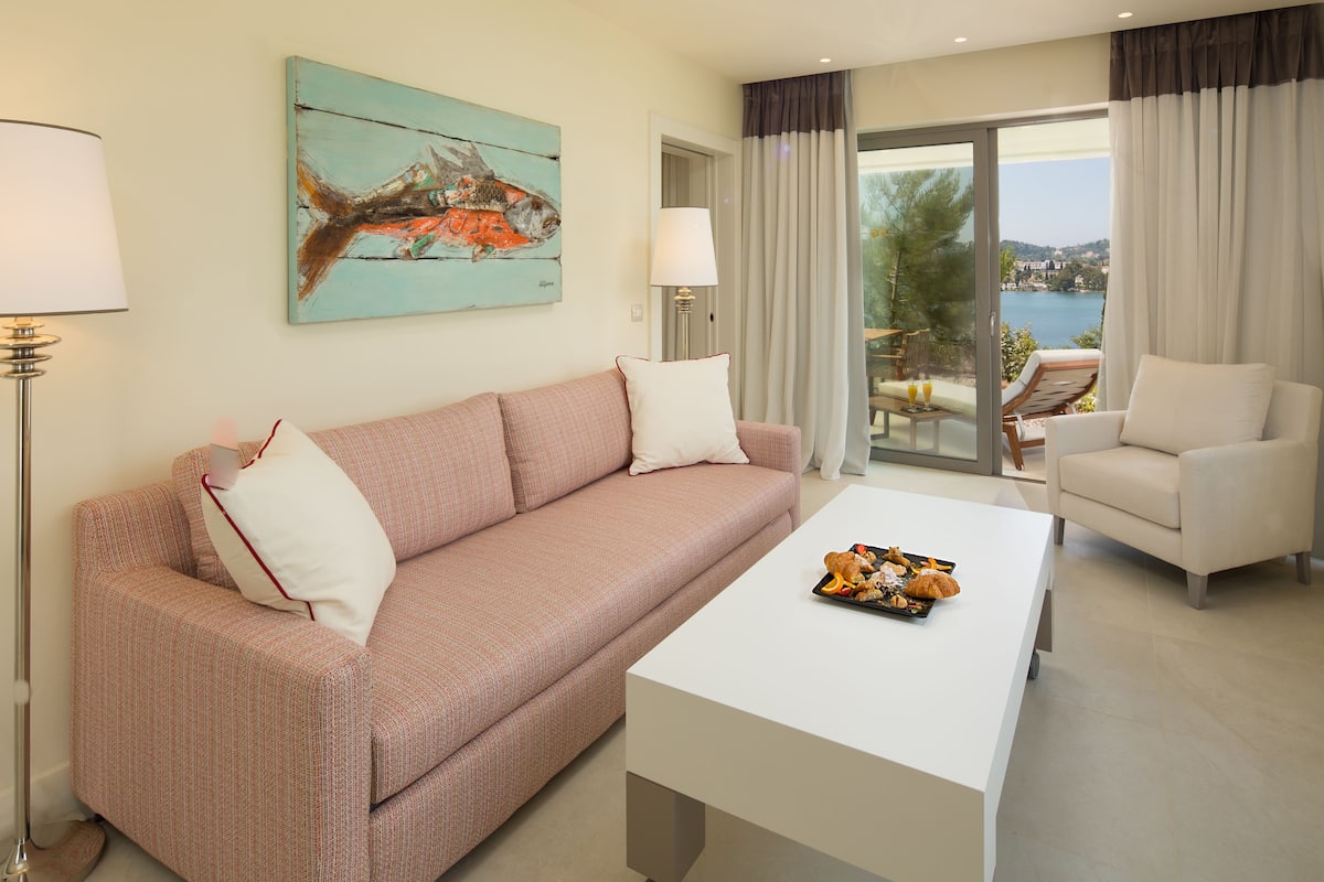 A modern living room with a sofa, a coffee table, and a chair. There is a painting on the wall and a view of the outdoors through the glass door.