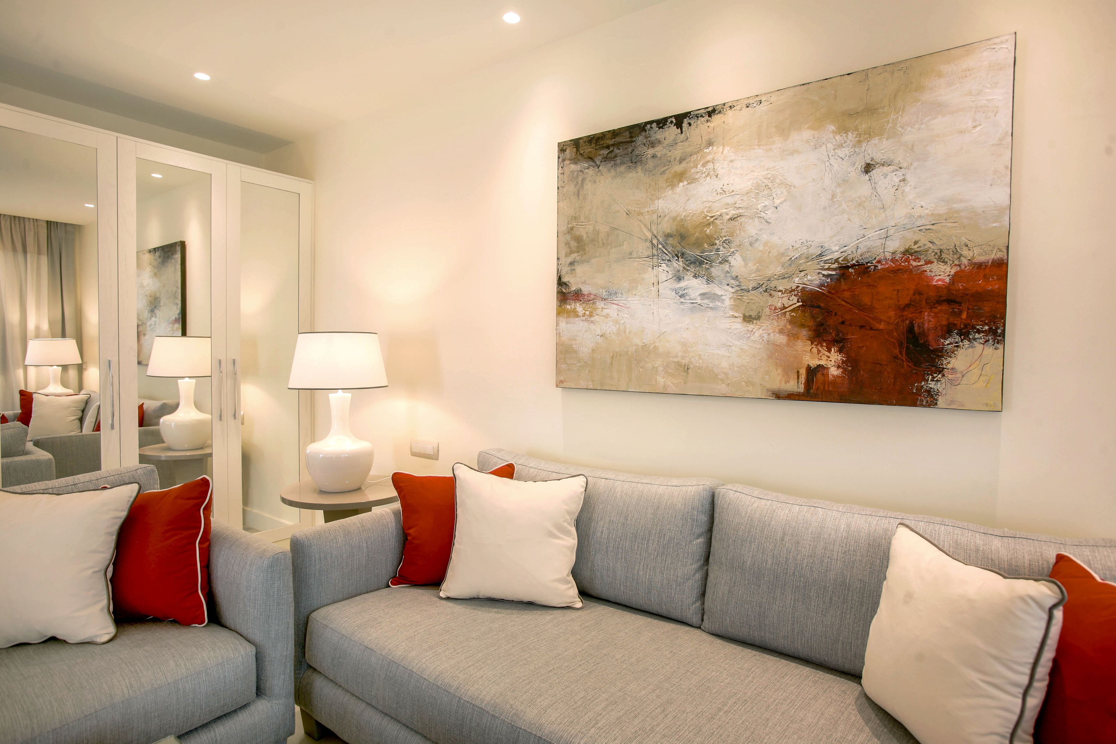 A modern living room with a grey sofa, white and red cushions, a large abstract painting, and a mirror reflecting the room.