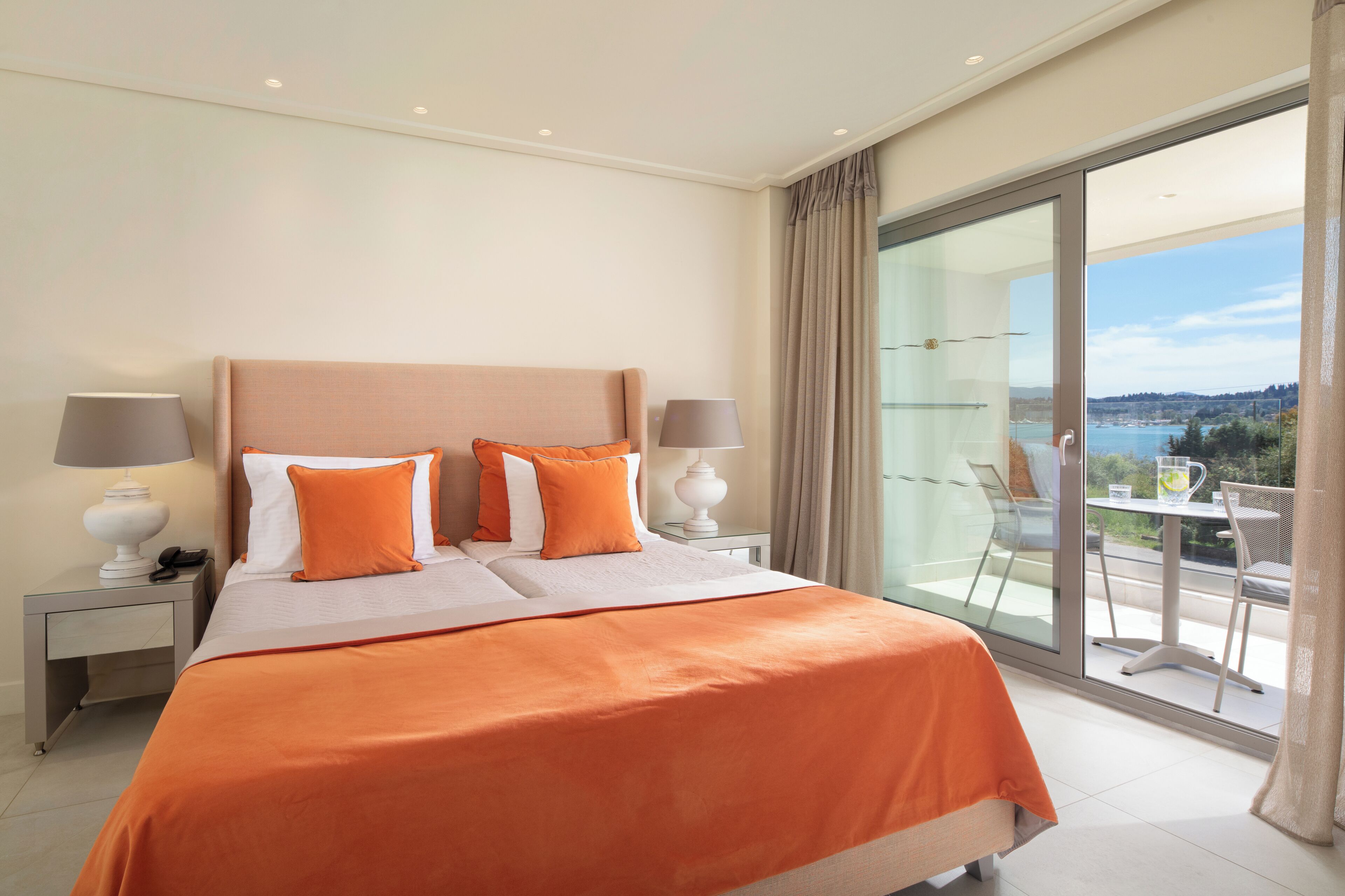 A hotel room with a large bed, two nightstands, a balcony with a table and chairs, and a view of the sea.
