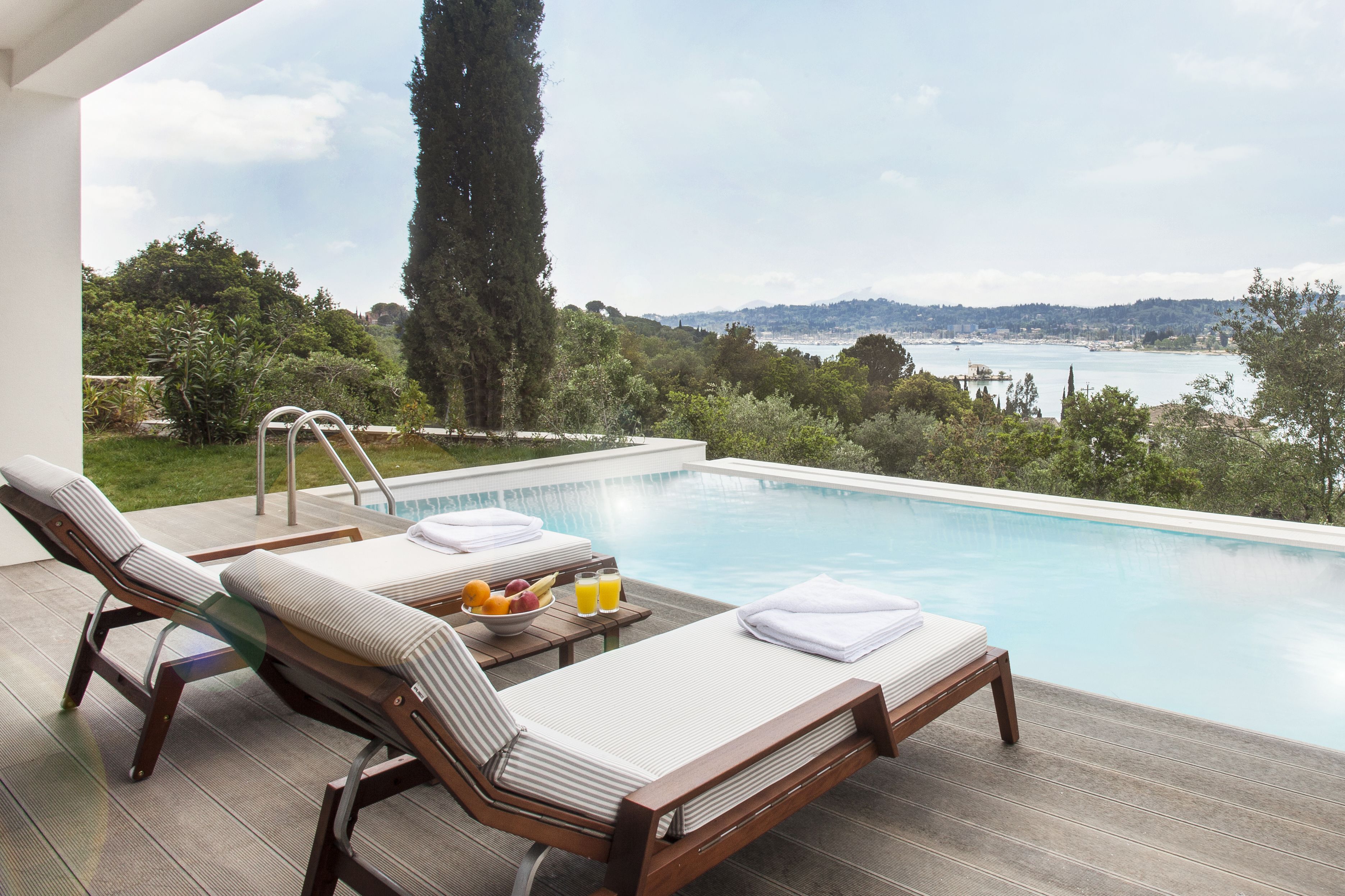 Un área junto a la piscina con sillones de descanso, una mesa con frutas y bebidas, y vistas a un cuerpo de agua y colinas.