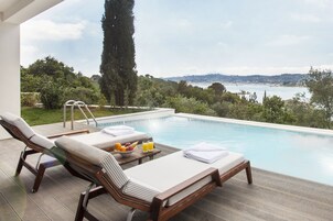 Un área junto a la piscina con sillones de descanso, una mesa con frutas y bebidas, y vistas a un cuerpo de agua y colinas.