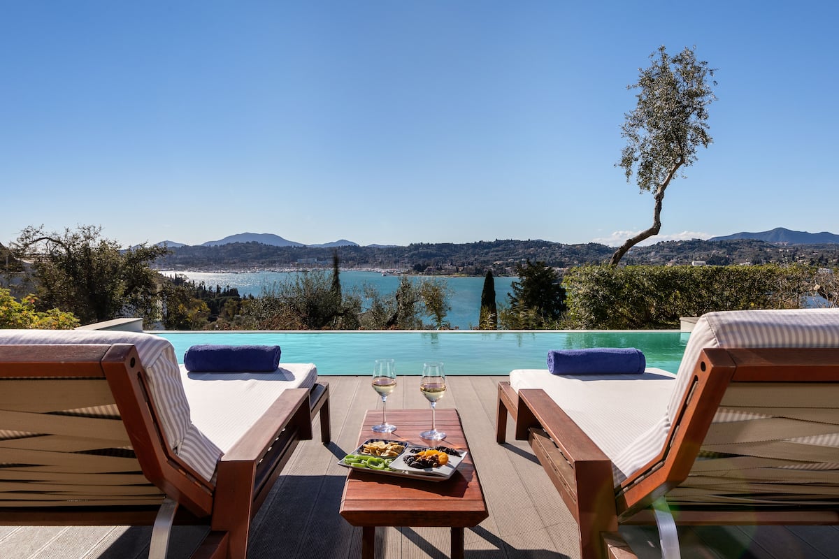 A poolside view with two lounge chairs, a table with a plate of food and two glasses of wine, and a clear view of the sea and mountains in the background.