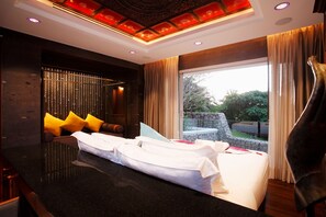 Habitación de hotel moderna con una cama grande, una zona de estar con cojines, una pared de piedra y vistas a zonas verdes.