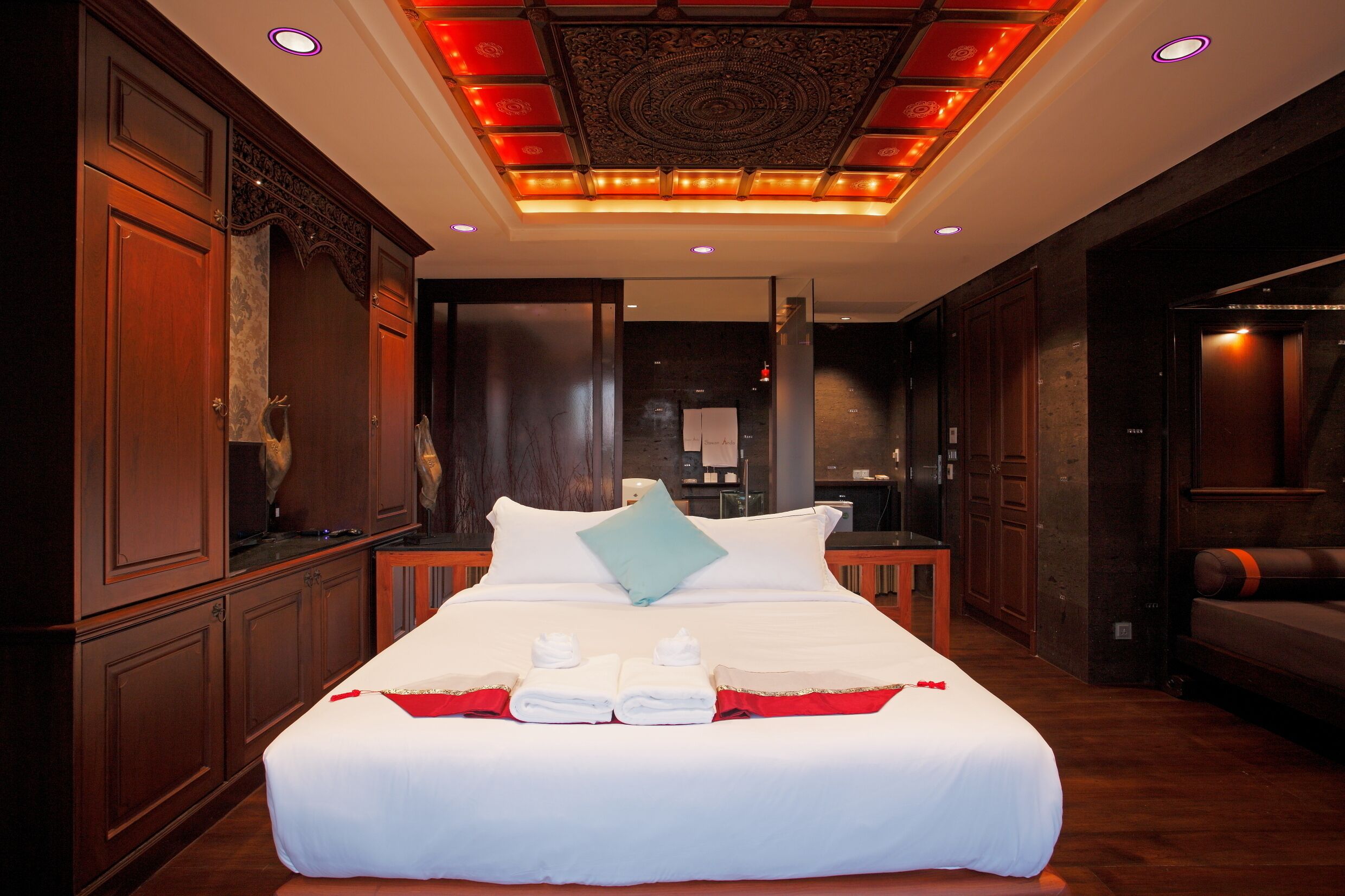 Una cama grande con sábanas blancas, un cojín decorativo rojo y dos toallas. La habitación tiene paneles de madera, un techo oscuro con un diseño circular e iluminación empotrada.