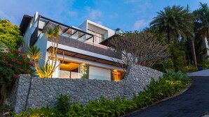Una casa moderna de dos pisos con un muro de piedra, abundante vegetación y un camino de entrada curvo.