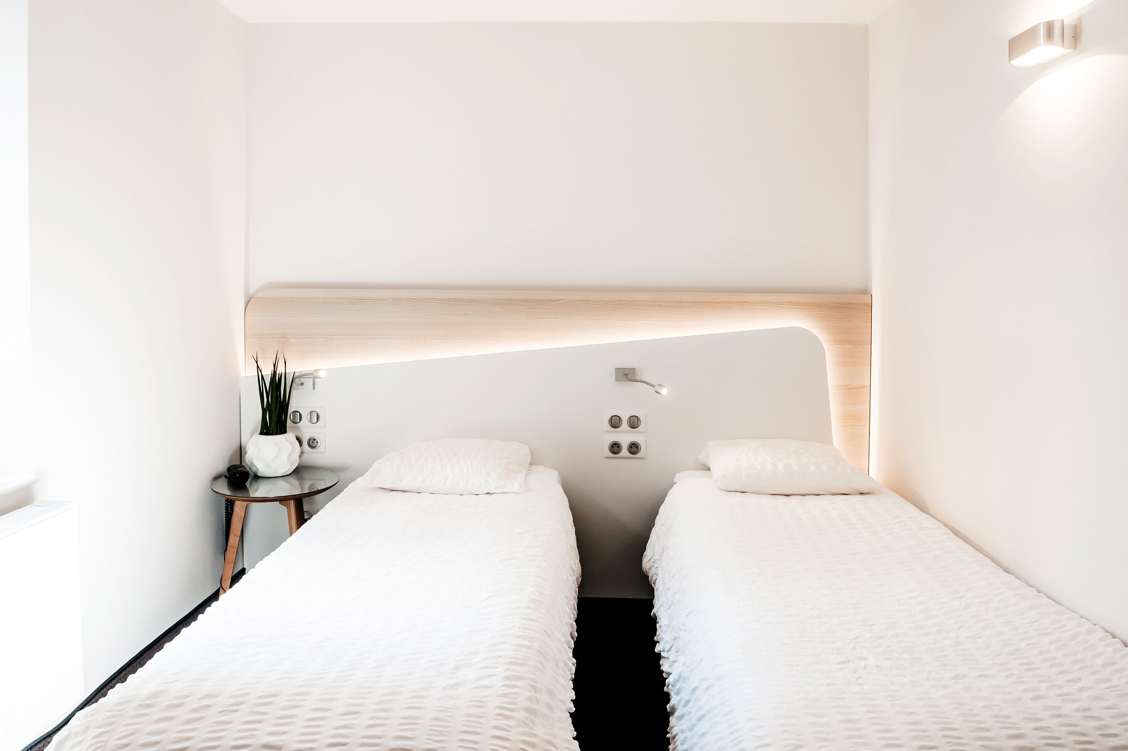 A minimalist hotel room with two beds, a small bedside table, a wall-mounted light, and a power outlet.