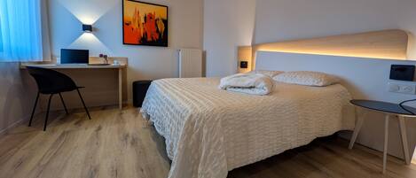 A hotel room with a bed, a desk, a chair, a radiator, and a painting on the wall.