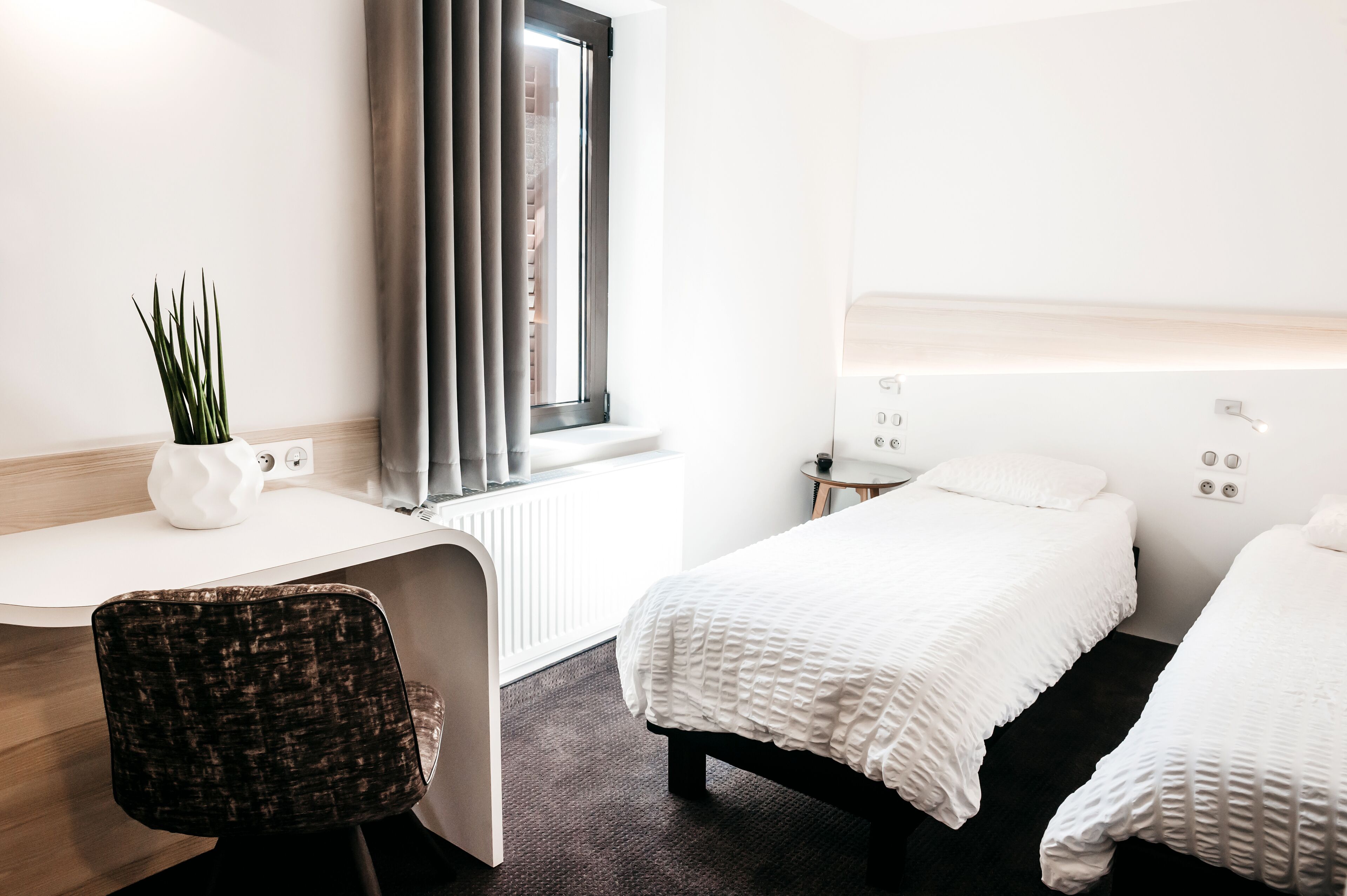 A hotel room with a desk, two beds, a potted plant, and a window with curtains.