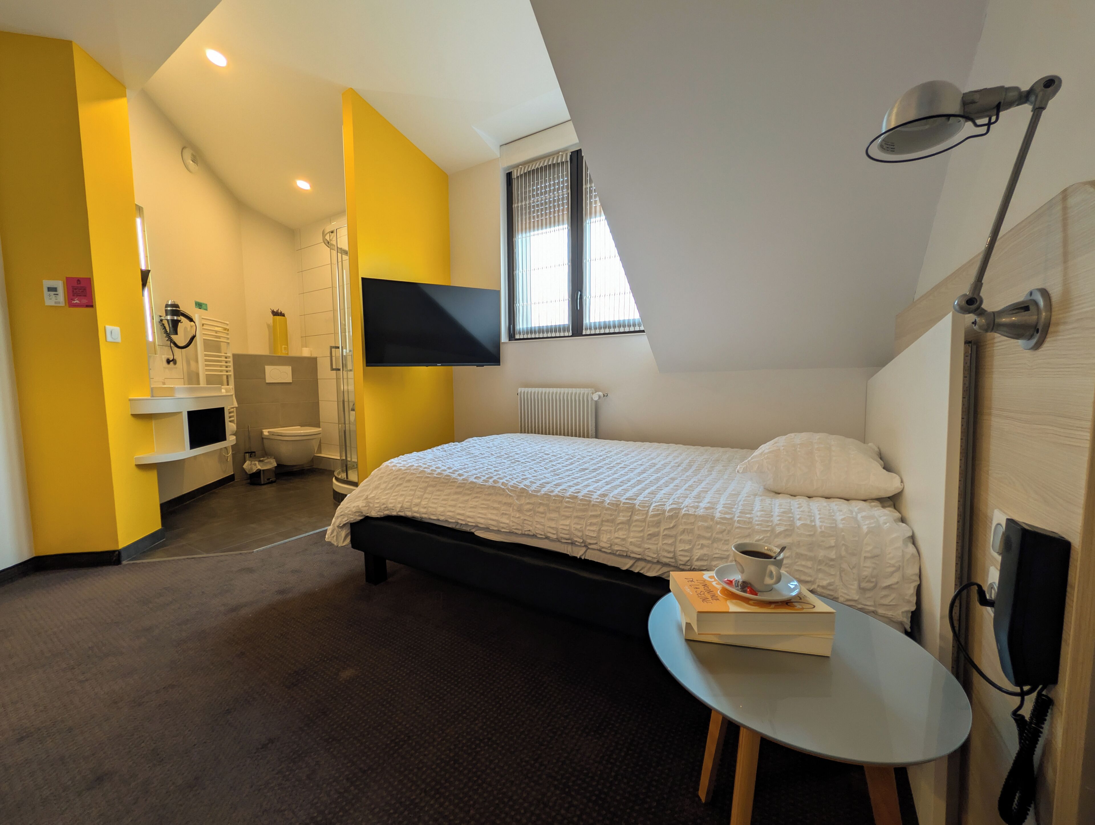 Een slaapkamer met een bed, televisie, raam, douche en een klein tafeltje met een boek en beker.