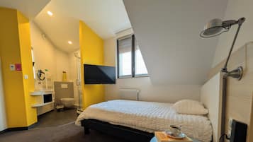 Een slaapkamer met een bed, televisie, raam, douche en een klein tafeltje met een boek en beker.