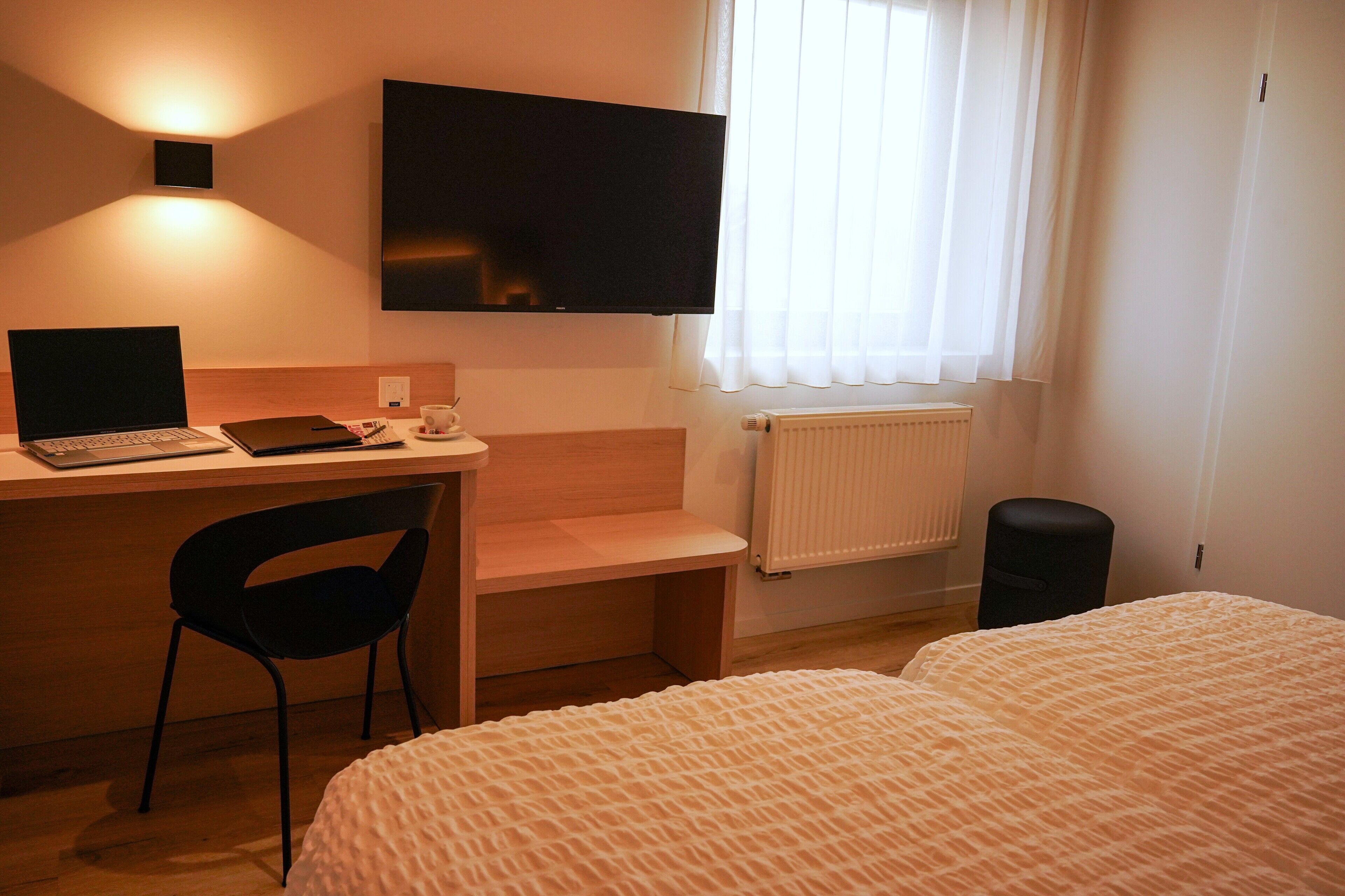 A hotel room with a desk, chair, bed, TV, and window with curtains.