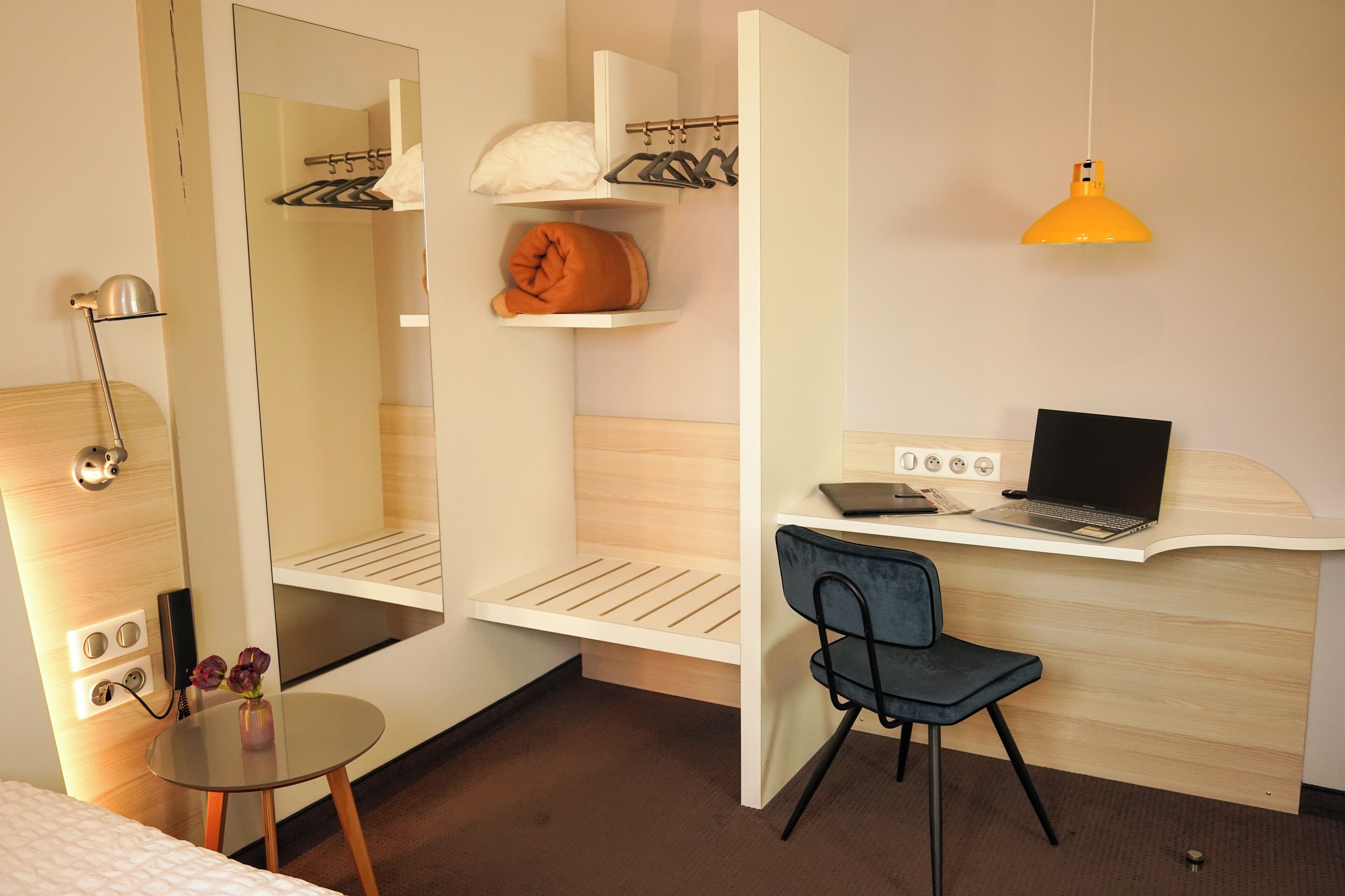 Habitación de hotel compacta con cama, escritorio con computadora portátil, silla, pequeña mesa con flores, espejo y lámpara colgante.