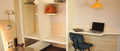 Habitación de hotel compacta con cama, escritorio con computadora portátil, silla, pequeña mesa con flores, espejo y lámpara colgante.