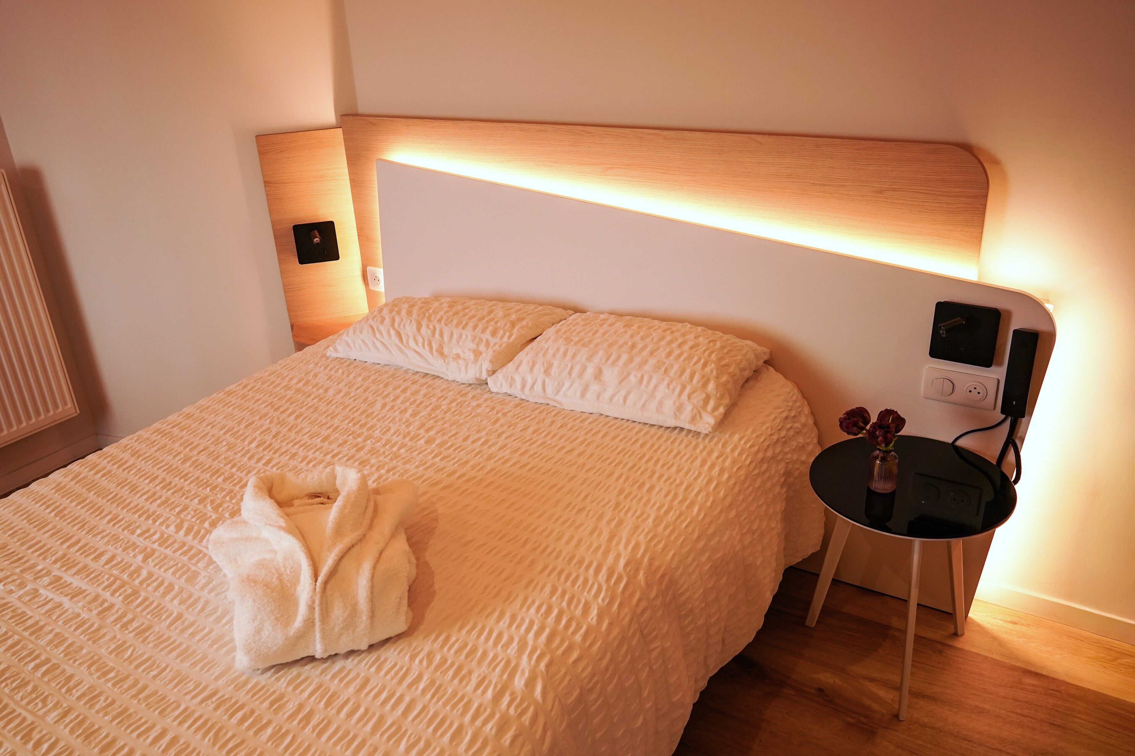 A neatly made bed with a white textured blanket and pillows, a small bedside table with a vase of flowers, and a wall-mounted control panel.
