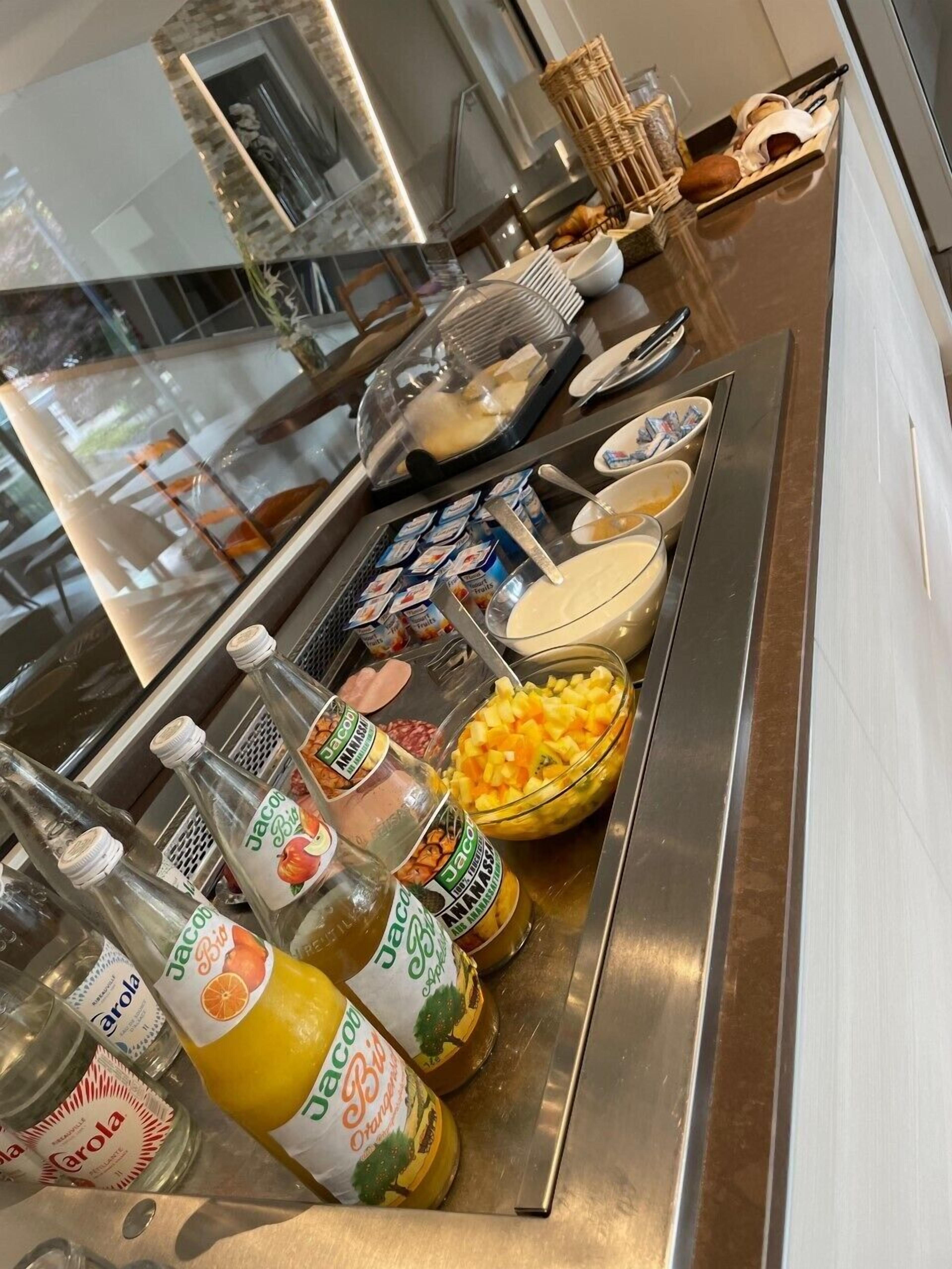 Un buffet con diversi cibi e bevande, tra cui bottiglie di Jacob's Bi e Carola, una ciotola di frutta a cubetti e un vassoio di pane a fette.