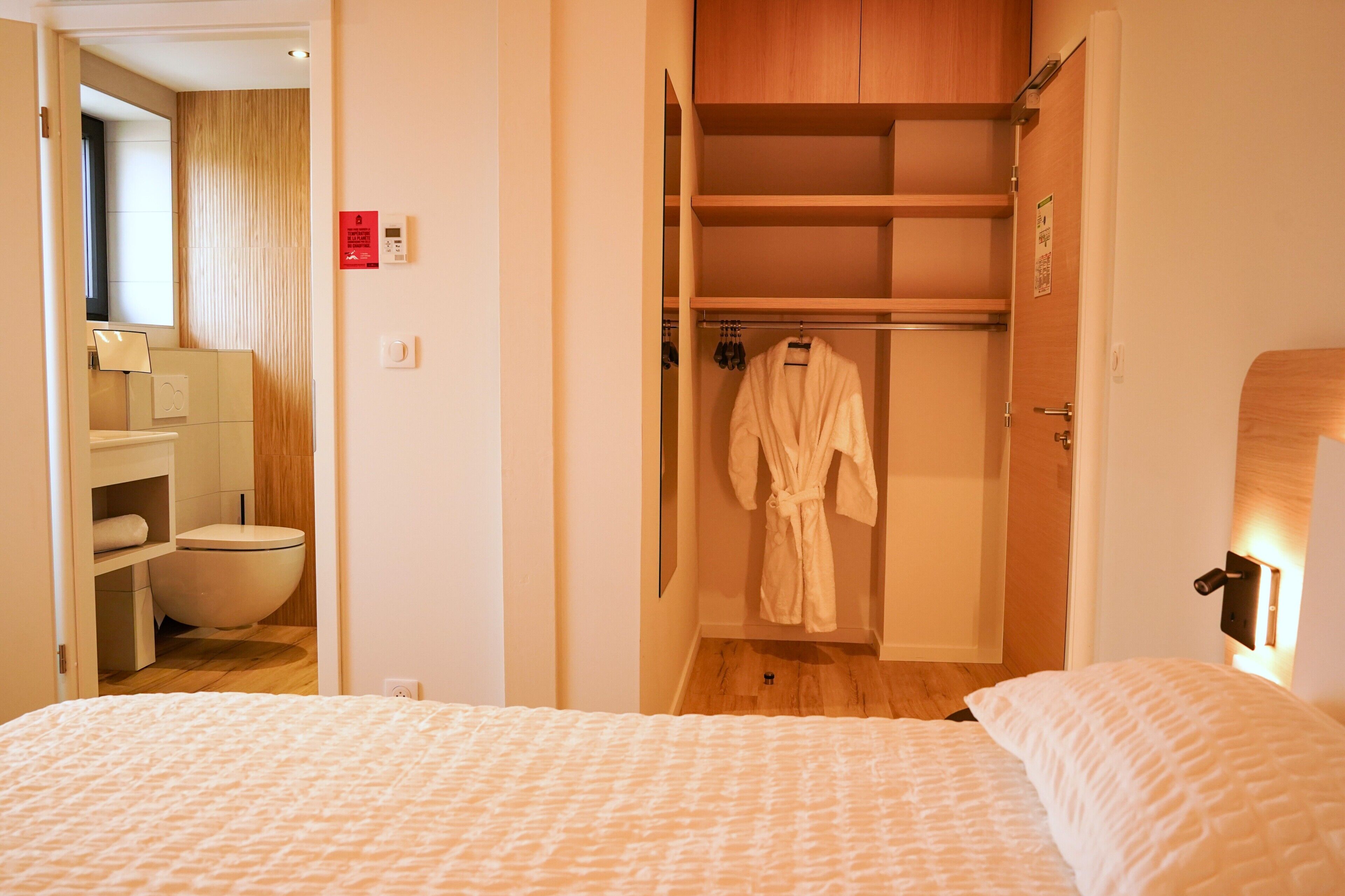A hotel room with a bed, a wooden wardrobe, a bathroom with a toilet, and a robe hanging inside the wardrobe.