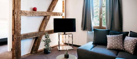 A living room with a wooden beam structure, a television on a stand, a sofa with patterned cushions, and a window with curtains.