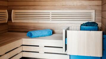 A sauna room with wooden benches, a blue towel, a wooden bucket with a white object, and a clock on the wall.