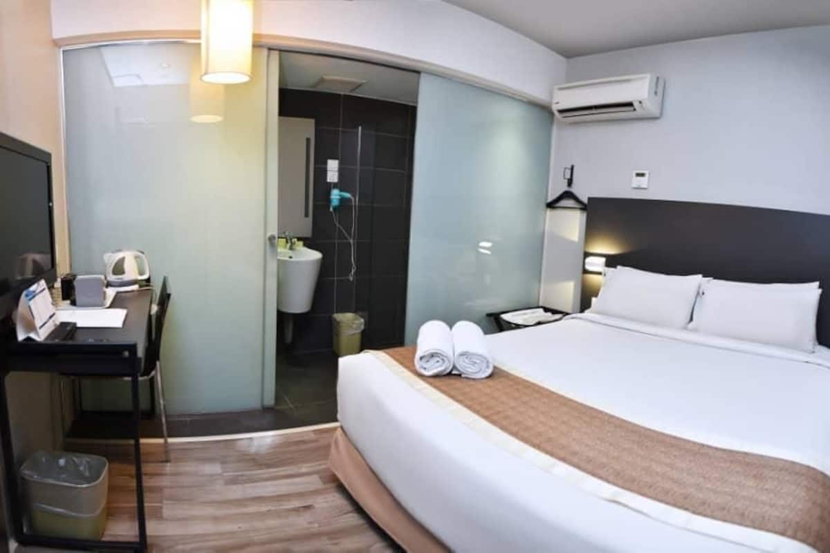 Habitación de hotel con cama, escritorio, silla, cesto de basura y zona de ducha.