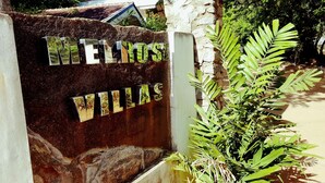 Een bord met de naam "MELIOS" en "VILLAS" op een rustieke stenen muur, omringd door groen.