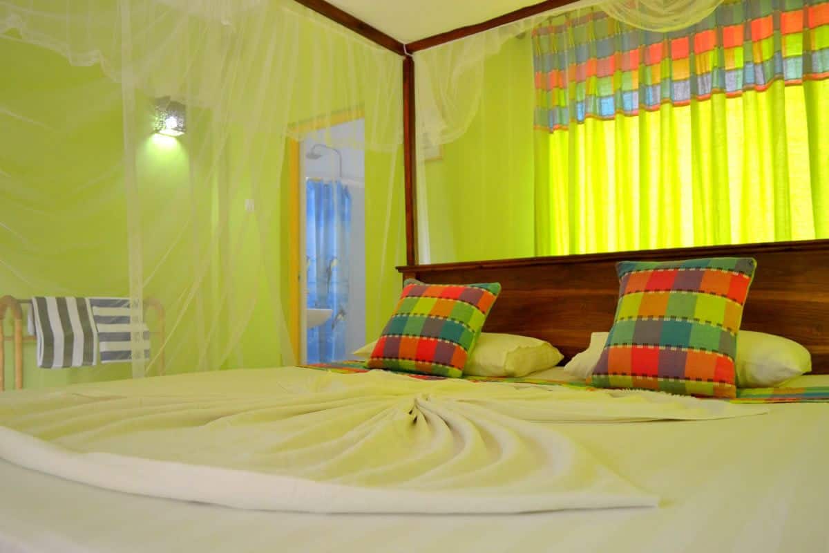Cama com travesseiros bem arrumados e colcha, cabeceira de madeira e cortinas leves.