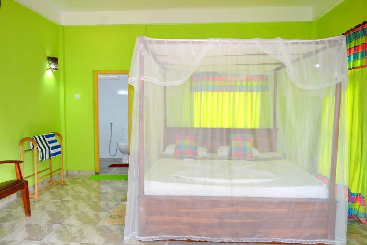 Quarto com cama sob mosquiteiro, um banco, uma porta e luminária fixada na parede.