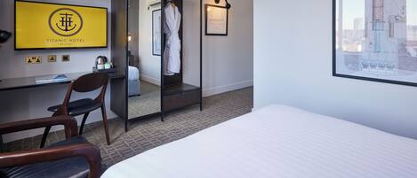 A hotel room with a bed, a desk with a computer, a chair, a robe hanging on a rack, and a framed picture of a building.
