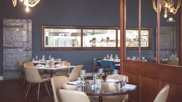 A restaurant with round tables set for dining, a stone wall, and a large window overlooking the kitchen.