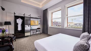 A hotel room with a bed, a desk with a lamp, a wardrobe, and a window with a view of the city.