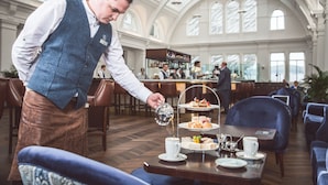 Un serveur apporte du thé et des pâtisseries dans une salle à manger spacieuse et bien éclairée, dotée de hauts plafonds et de grandes fenêtres.