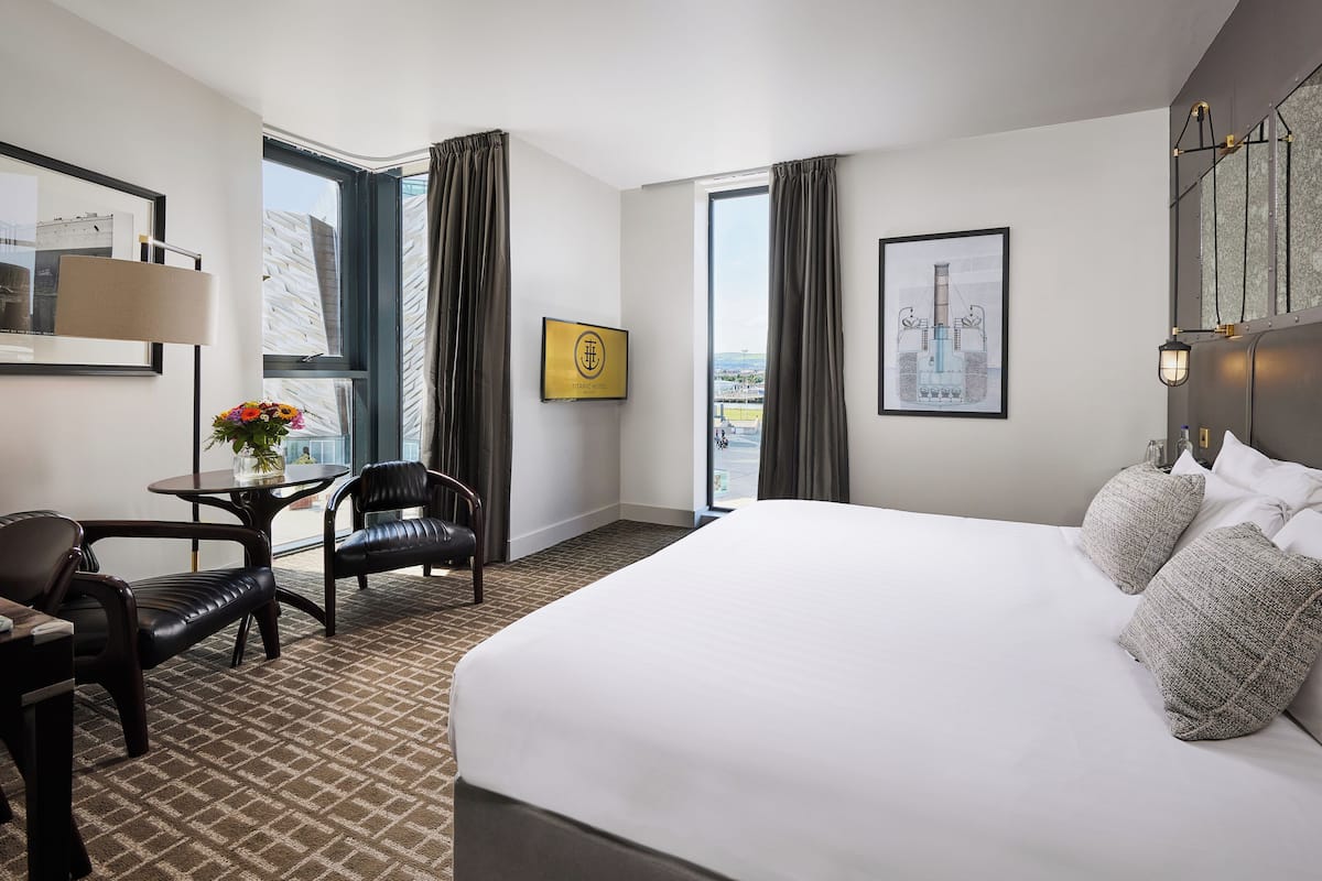 Habitación de hotel con una cama grande, un escritorio con silla, una mesita con un jarrón de flores y vistas a la ciudad a través de amplias ventanas.
