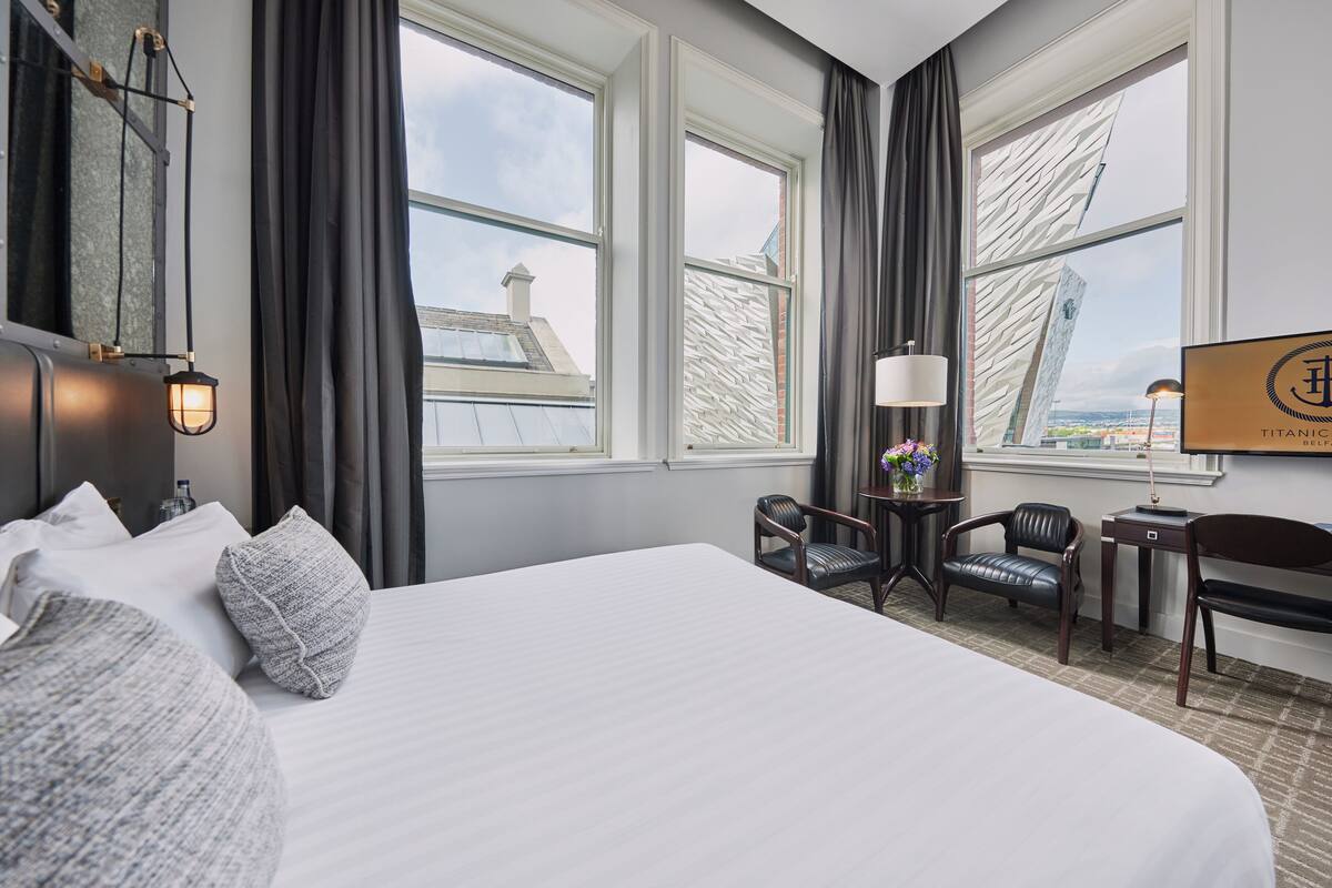 Habitación de hotel con una cama grande, dos sillas, una mesita y vistas a un edificio a través de las ventanas.