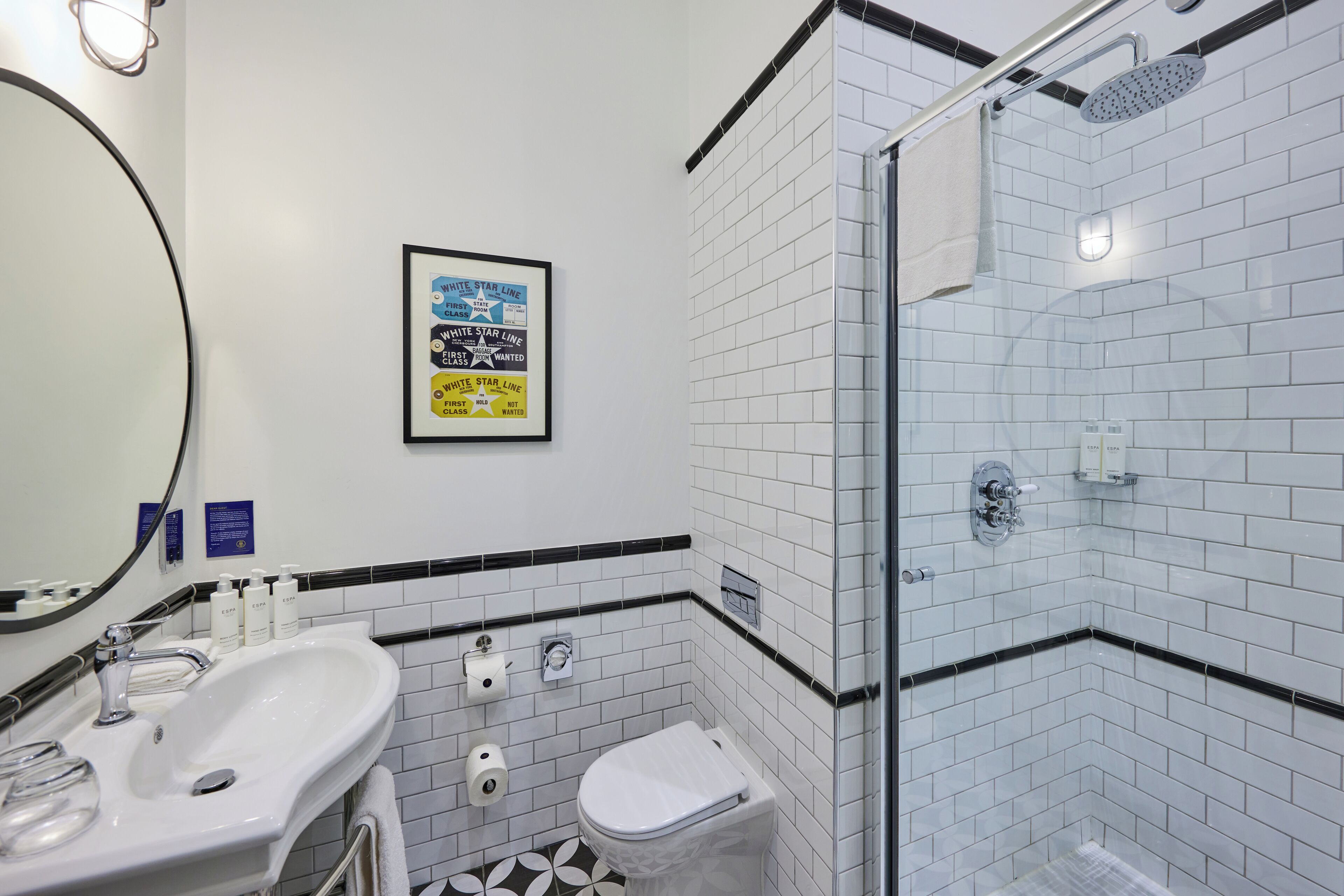 Un baño moderno con lavamanos, inodoro y ducha blancos. Hay un cartel enmarcado en la pared y una toalla colgada en la puerta de la ducha.