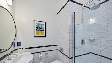 Un baño moderno con lavamanos, inodoro y ducha blancos. Hay un cartel enmarcado en la pared y una toalla colgada en la puerta de la ducha.