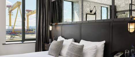 A hotel room with a large bed, two pillows, a nightstand with a phone and water bottle, a mirror, and a view of construction cranes outside.