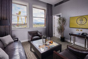 Et moderne hotelværelse med en lædersofa, et glasbord med champagne, glas og en skål med frugt. Der er et stort vindue med udsigt til en byggeplads og en dør, der fører til et andet værelse.