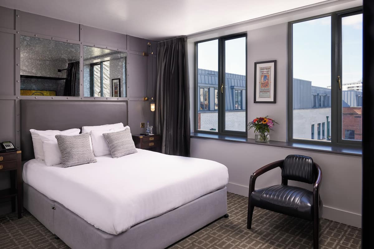 Habitación de hotel con una cama grande, una silla, una mesita de noche, un espejo y una ventana con vistas a edificios.