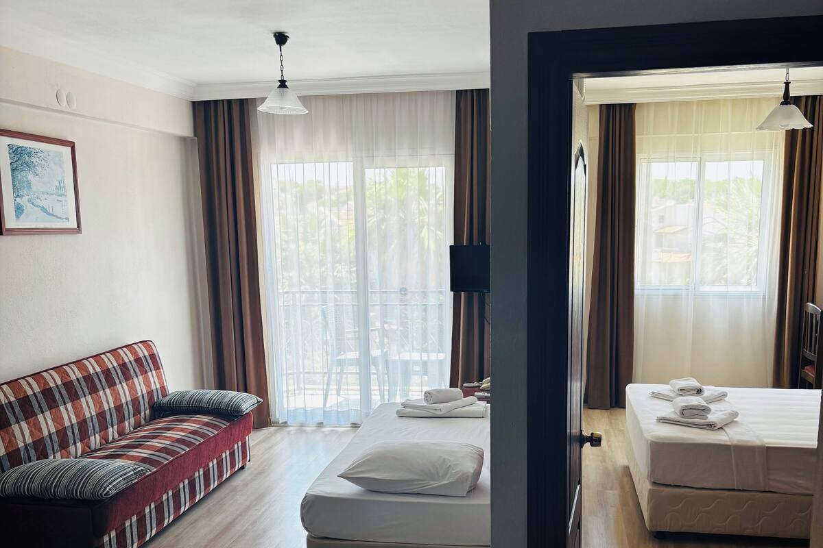 Quarto de hotel com sofá, cama, televisão e varanda com cortinas.