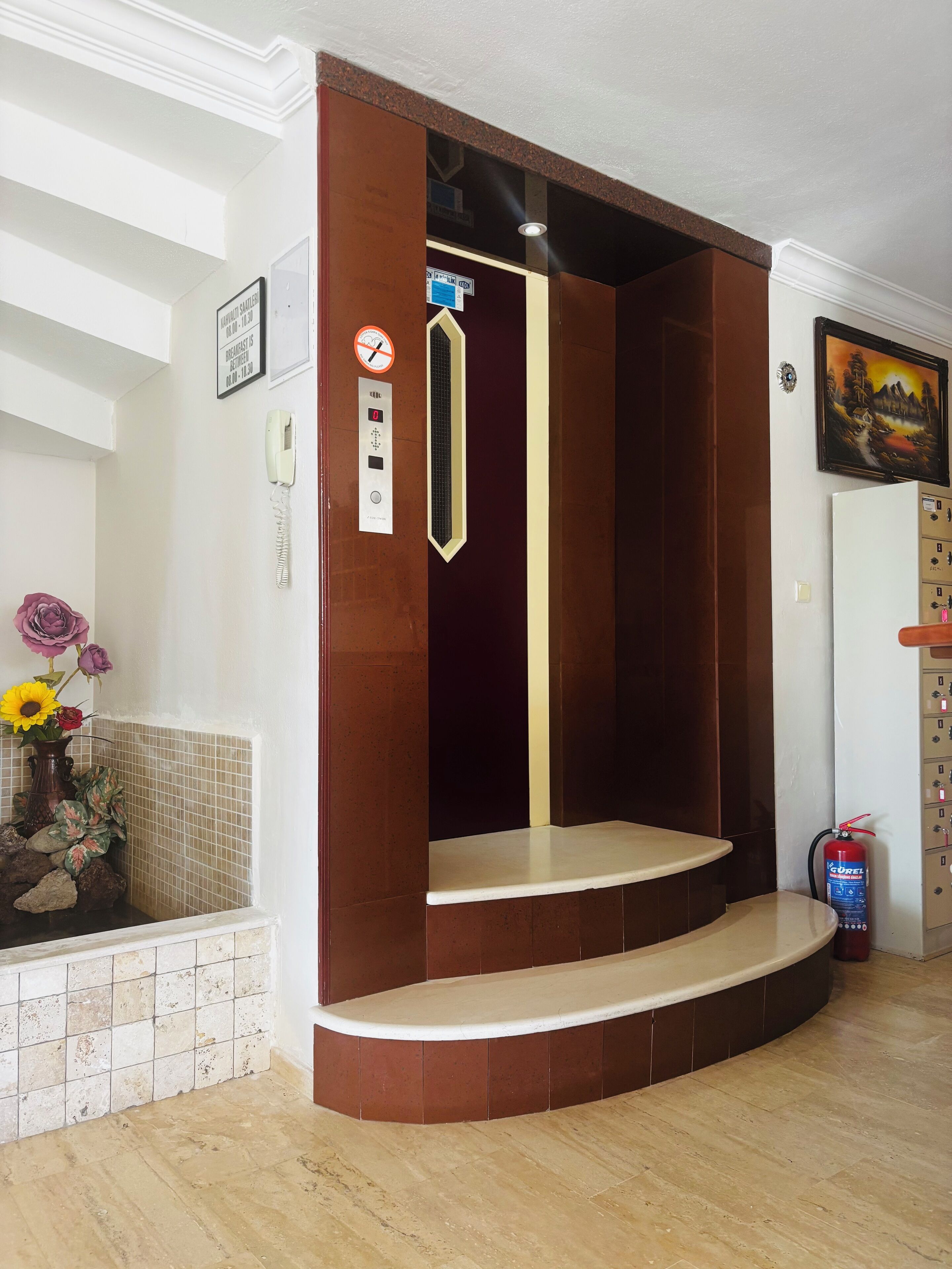 Een hotel lift met een bedieningspaneel, een brandblusser en een decoratieve vaas met bloemen.