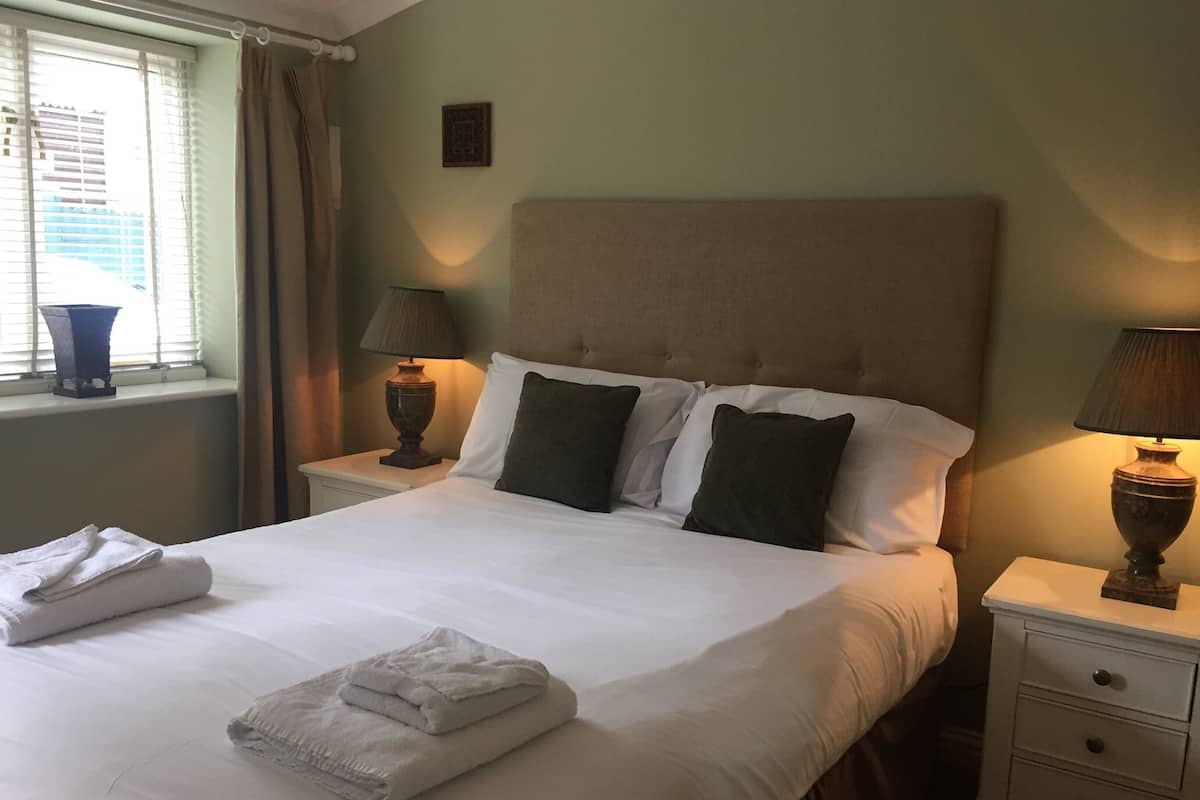 Cama bem arrumada com lençóis brancos, duas luminárias de cabeceira e uma janela com cortinas.