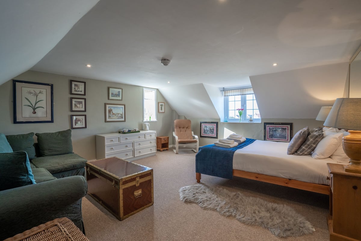 Um quarto com teto estilo loft, uma cama com manta azul, cômoda, cadeira e quadros nas paredes.