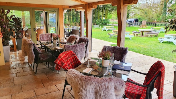 An outdoor patio with tables and chairs, covered by a wooden structure, overlooking a grassy area with picnic tables. - Bedingfeld Arms (King's Lynn)