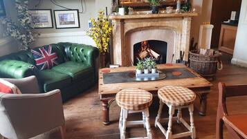 A cozy living room with a green leather sofa, a wooden coffee table with stools, a fireplace, and a wall adorned with framed pictures and a potted plant.