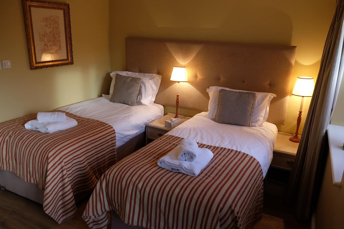 Quarto de hotel com duas camas de solteiro, cada uma com colcha listrada, travesseiros e toalha dobrada. Há luminárias de cabeceira, mesas de apoio e um quadro na parede.