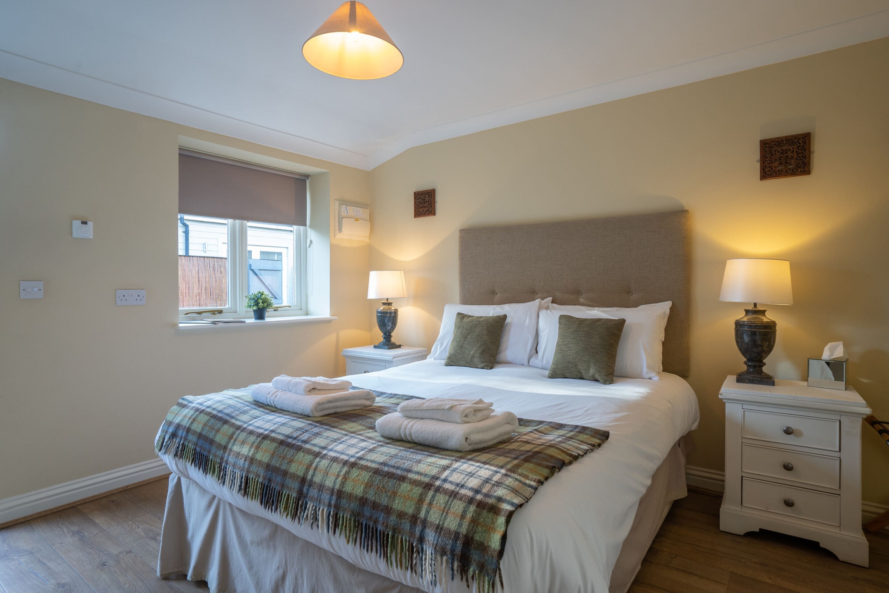 Een keurig opgemaakt bed met een geruit plaid, twee bedlampen en een raam met jaloezieën.