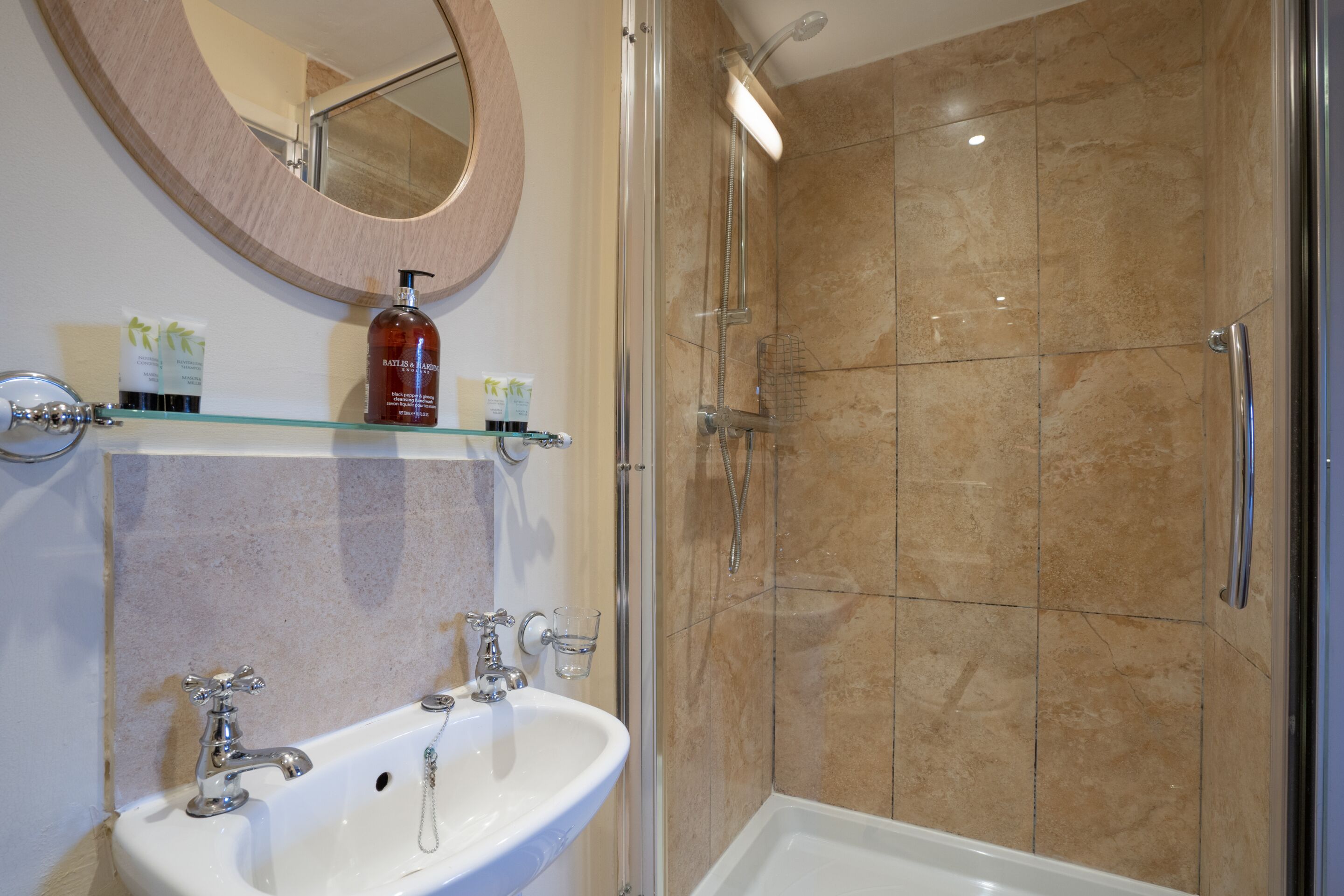A bathroom with a glass-enclosed shower, a white sink with two faucets, and a round mirror above the sink.
