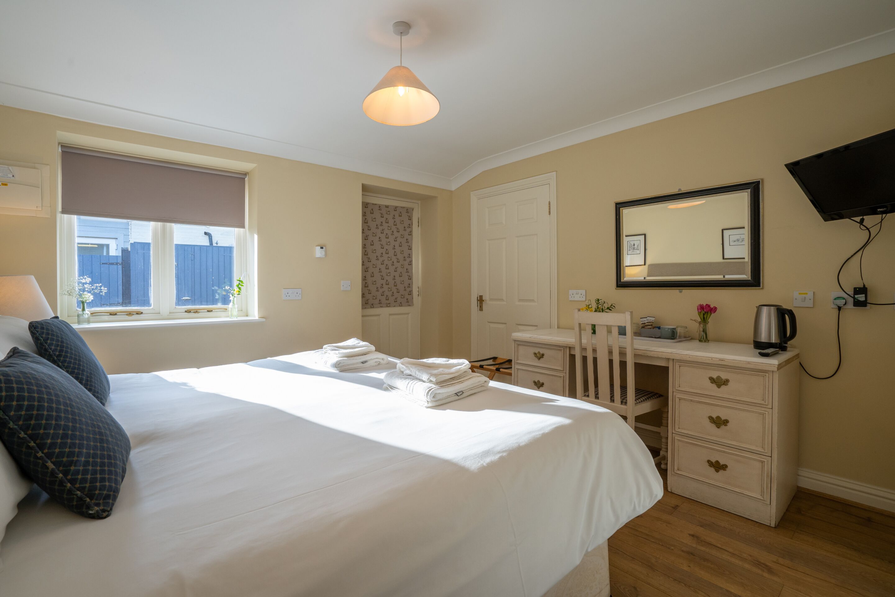 A hotel room with a bed, a desk, a chair, a dresser, a TV, and a window with a view of a blue fence.