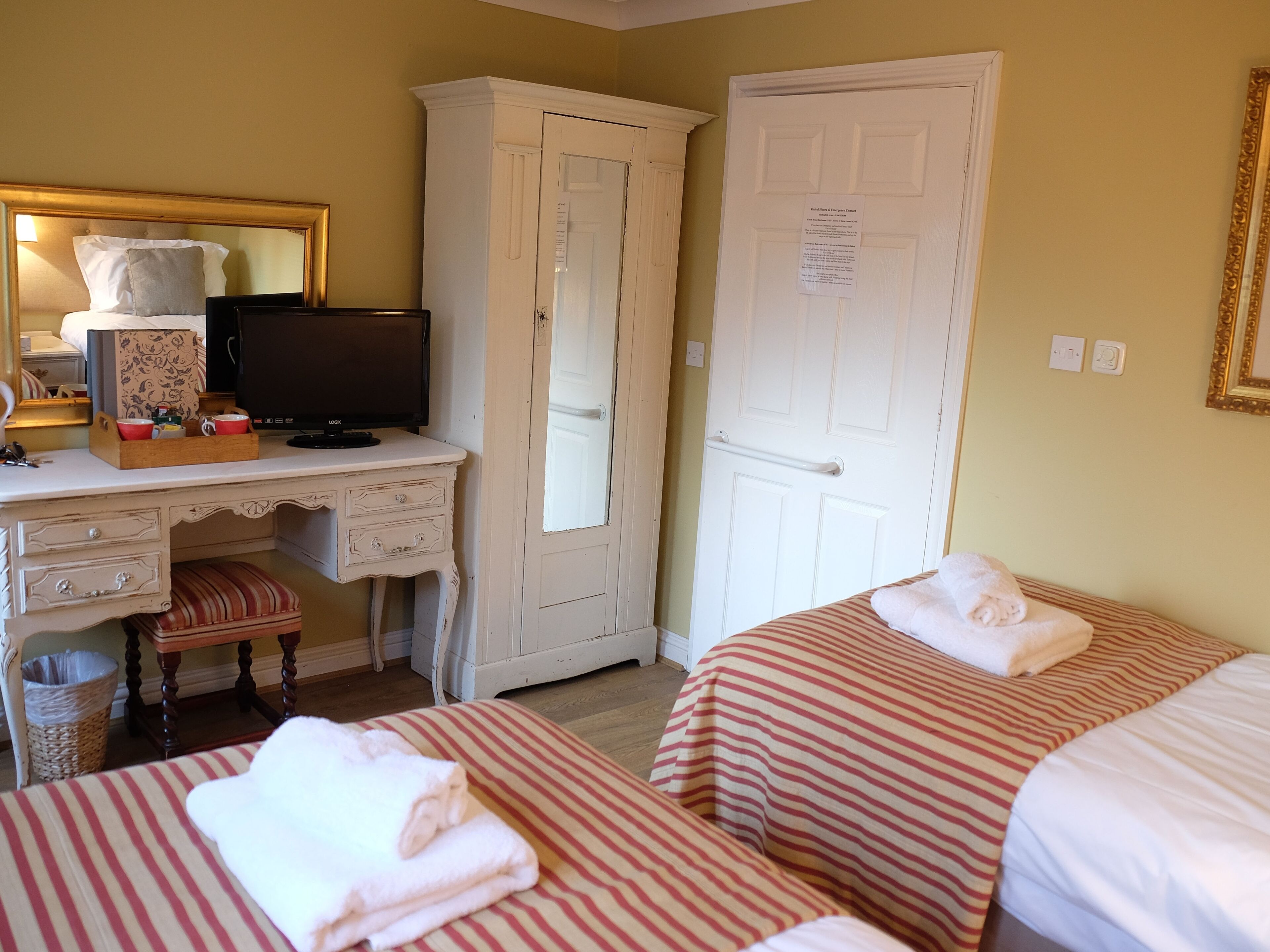 A hotel room with a desk, television, wardrobe, and two beds with striped bedding.